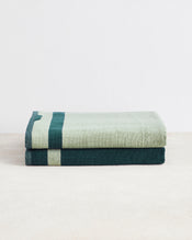 The Bed Threads Forest & Sage Two-Toned 100% Cotton Terry Bath Sheets set features two plush, quick-drying towels—one light green with a dark green stripe, one dark green with a light green stripe—stacked on a light surface against a white background. - view