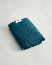 The Aegean 100% Cotton Terry Bath Towel by Bed Threads, a teal 650GSM towel with a small white tag, rests on a light gray textured surface and offers quick-drying convenience. - view