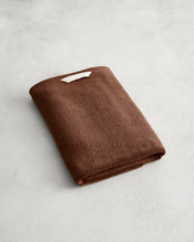 The Bed Threads Espresso 100% Cotton Terry Bath Towel, neatly folded with a small white label, rests on a light gray textured surface. - view