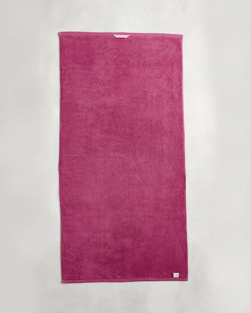 A rectangular Berry 100% Cotton Terry Bath Towel by Bed Threads hangs flat against a light gray wall. The plush magenta towel has a small white tag on the bottom right corner.