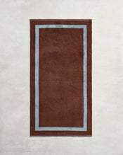 The Cacao & Lagoon Two-Toned 100% Cotton Terry Bath Towel by Bed Threads adds a cozy touch when paired with a brown rug featuring a light blue border, creating an inviting vibe for your bathroom. - view