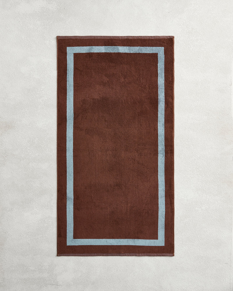 The Cacao & Lagoon Two-Toned 100% Cotton Terry Bath Towel by Bed Threads adds a cozy touch when paired with a brown rug featuring a light blue border, creating an inviting vibe for your bathroom.