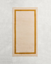 The Bed Threads Crème & Turmeric Two-Toned 100% Cotton Terry Bath Towel, a beige rectangular 650GSM towel with a thick mustard yellow border, is shown laid flat on a light textured surface. - view
