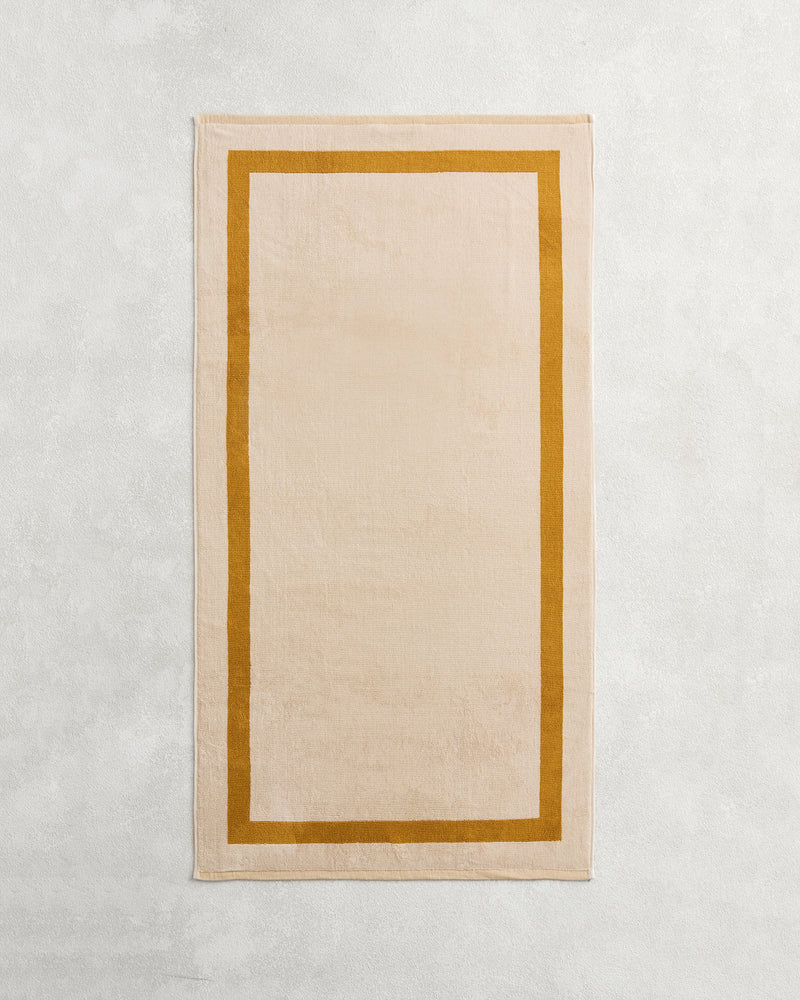 The Bed Threads Crème & Turmeric Two-Toned 100% Cotton Terry Bath Towel, a beige rectangular 650GSM towel with a thick mustard yellow border, is shown laid flat on a light textured surface.
