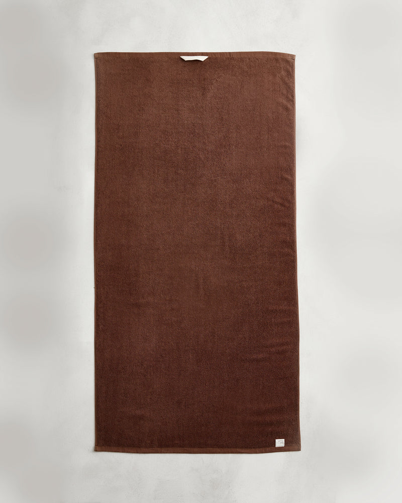 An Espresso 100% Cotton Terry Bath Towel by Bed Threads is laid flat on a light gray surface, featuring a small white label on the bottom corner and a beige hanging loop near the top edge.