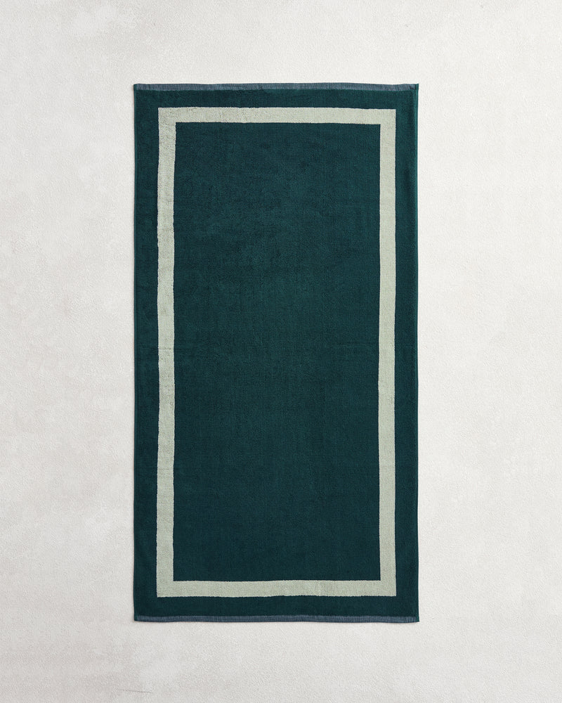 A rectangular dark green rug with a beige double border, set on a light surface, creates a serene space—perfect for pairing with the Forest & Sage Two-Toned 100% Cotton Terry Bath Towel Bundle from Bed Threads.