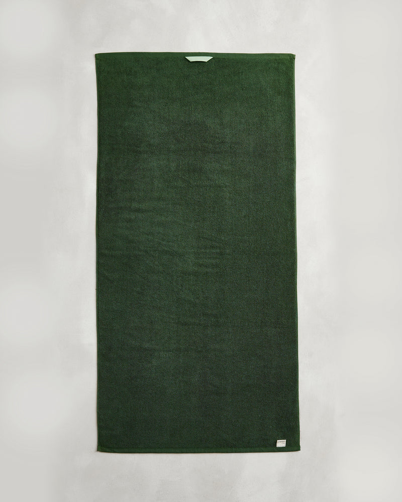 A Moss 100% Cotton Terry Bath Towel from Bed Threads hangs vertically against a white background, displaying a light blue tag at the top and a small label in the bottom right corner.