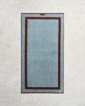 The Bed Threads Cacao & Lagoon Two-Toned 100% Cotton Terry Bath Towel Bundle is shown laid flat, featuring a blue rectangular center, dark brown border with an inner brown frame, and a small tag on the bottom right corner. - view