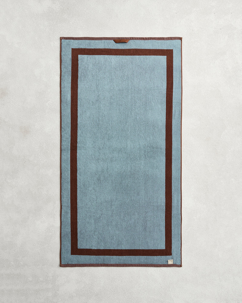 The Bed Threads Cacao & Lagoon Two-Toned 100% Cotton Terry Bath Towel Bundle is shown laid flat, featuring a blue rectangular center, dark brown border with an inner brown frame, and a small tag on the bottom right corner.