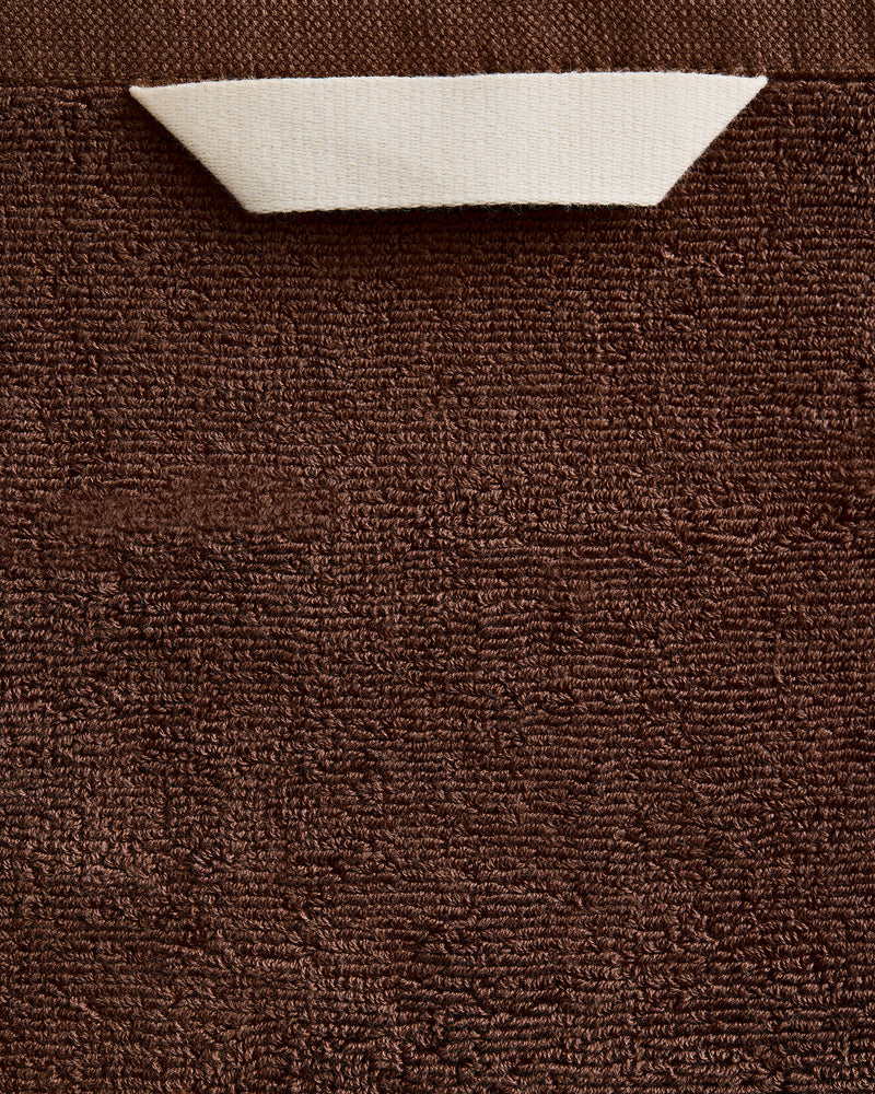 A close-up of the Bed Threads Espresso 100% Cotton Terry Bath Towel, showing its brown textured surface with a white triangular corner folded down to reveal the reverse side.