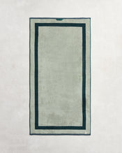 The Bed Threads Forest & Sage Two-Toned 100% Cotton Terry Bath Towel, in light gray with a dark green border and deeper green inner outline, is shown laid flat on a light textured surface. - view