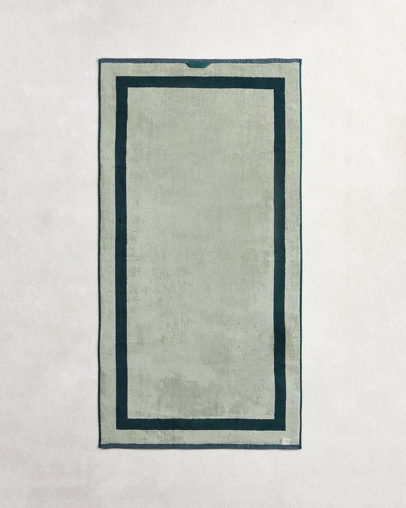 The Bed Threads Forest & Sage Two-Toned 100% Cotton Terry Bath Towel, in light gray with a dark green border and deeper green inner outline, is shown laid flat on a light textured surface.