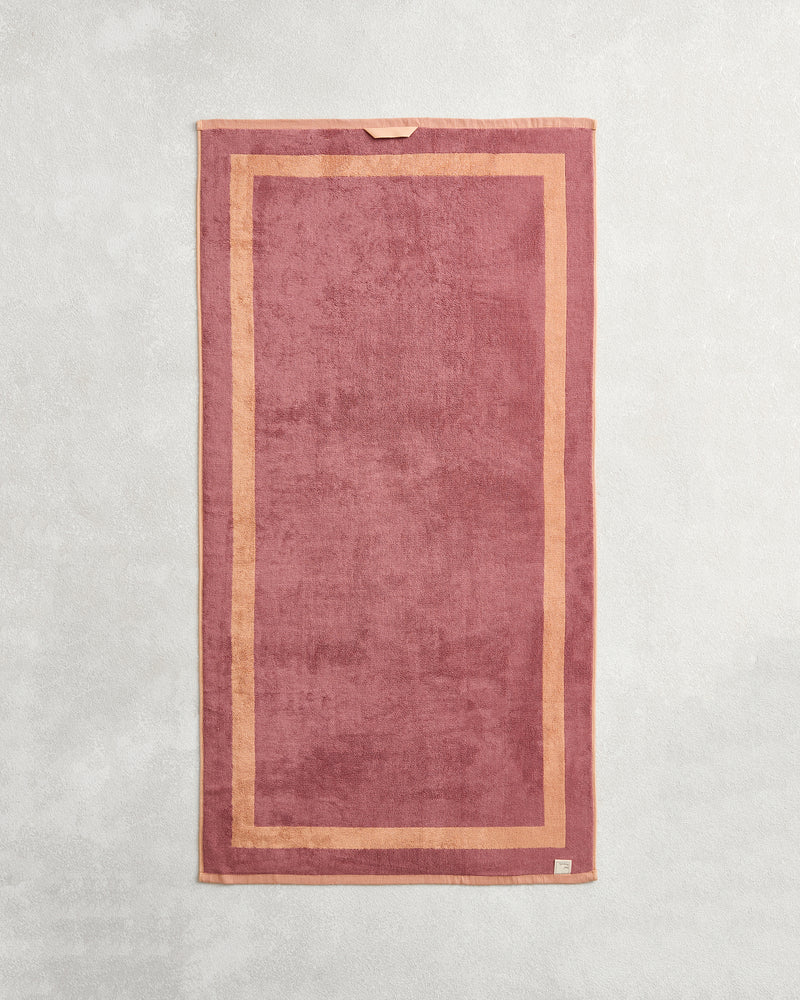 The Bed Threads Terracotta & Pink Clay Two-Toned 100% Cotton Terry Bath Towel Bundle features rectangular bath towels in rose and light pink, with a subtle border and beige tag, displayed flat on a light, textured surface.