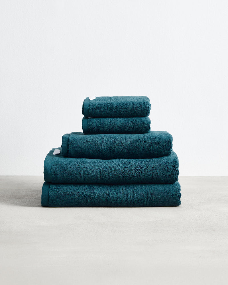 A stack of four Aegean 100% Cotton Terry Bath Towels by Bed Threads in dark teal rests neatly folded on a light surface against a plain white background.