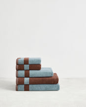 Four neatly folded Cacao & Lagoon Two-Toned 100% Cotton Terry Bath Towels from Bed Threads are stacked on a light surface. The bundle features towels with brown stripes and trim, arranged largest to smallest for a stylish look. - view