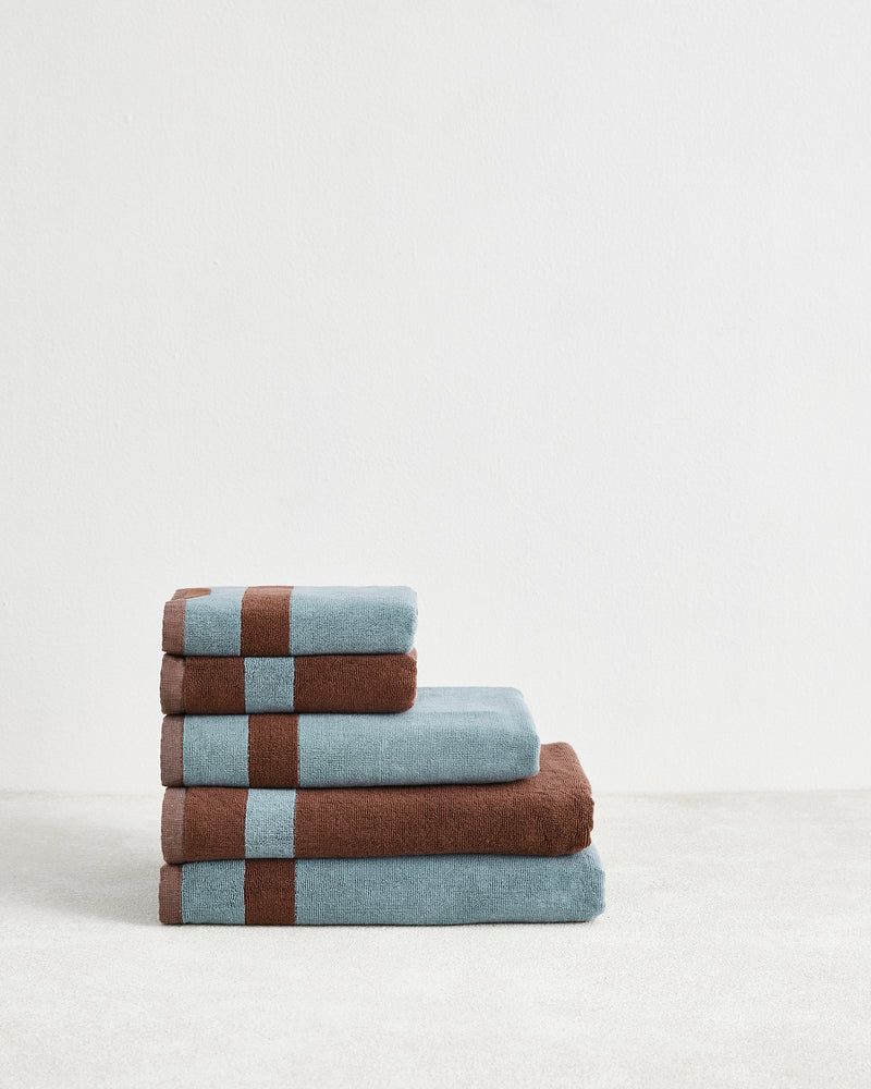 Four neatly folded Cacao & Lagoon Two-Toned 100% Cotton Terry Bath Towels from Bed Threads are stacked on a light surface. The bundle features towels with brown stripes and trim, arranged largest to smallest for a stylish look.