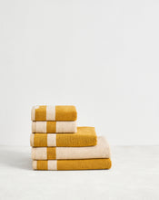 The Bed Threads Crème & Turmeric Two-Toned 100% Cotton Terry Bath Towel Bundle features four plush towels with a white stripe, neatly stacked on a light surface against a plain white wall. - view