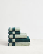 A neatly folded stack of four Forest & Sage Two-Toned 100% Cotton Terry Bath Towels by Bed Threads, arranged from largest to smallest, sits on a light surface with a white background. - view