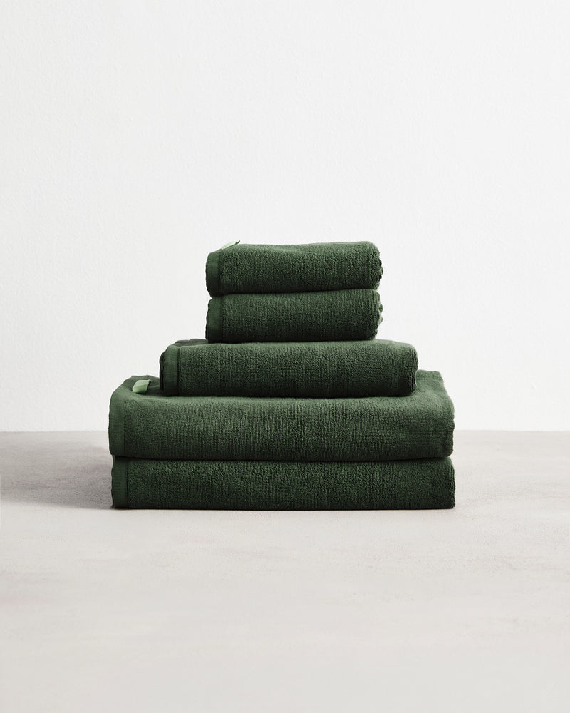 A neatly stacked Bed Threads Moss 100% Cotton Terry Bath Towel Bundle in dark green, featuring various sizes, is arranged on a light gray surface against a plain white background.