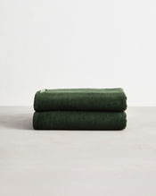 Two Moss 100% Cotton Terry Bath Towels (Set of Two) by Bed Threads, neatly folded in dark green, are stacked on a light surface with a plain white background. - view