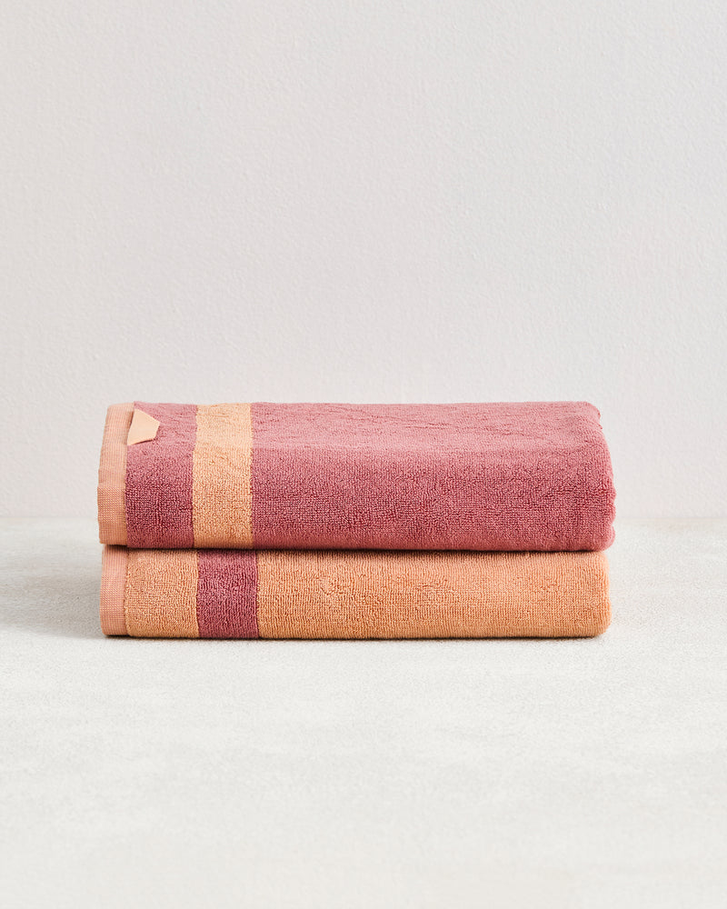 A set of two Bed Threads Terracotta & Pink Clay Two-Toned 100% Cotton Terry Bath Towels, one with a gold stripe and the other with a pink stripe, neatly folded on a light surface against a pale background.