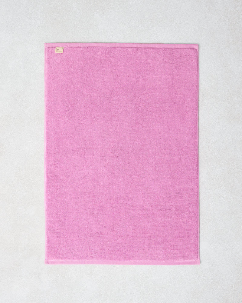 The Bubblegum 100% Cotton Terry Bath Mat by Bed Threads, in a rectangular pink design, lies flat on a light textured surface with a small fabric tag visible on one corner.