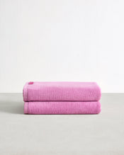A set of two Bubblegum 100% Cotton Terry Bath Towels by Bed Threads, neatly folded and highly absorbent, are stacked on a light gray surface against a plain white background. - view
