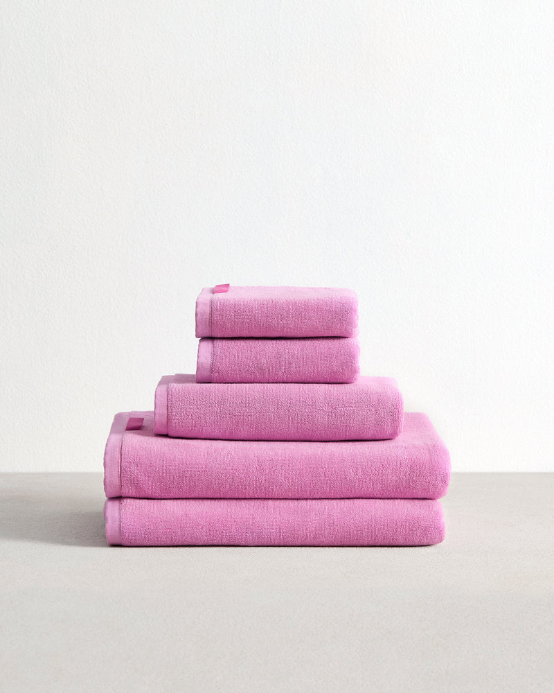 The Bed Threads Bubblegum 100% Cotton Terry Bath Sheet Bundle features four absorbent pink towels in assorted sizes, neatly stacked on a light surface against a plain white background.