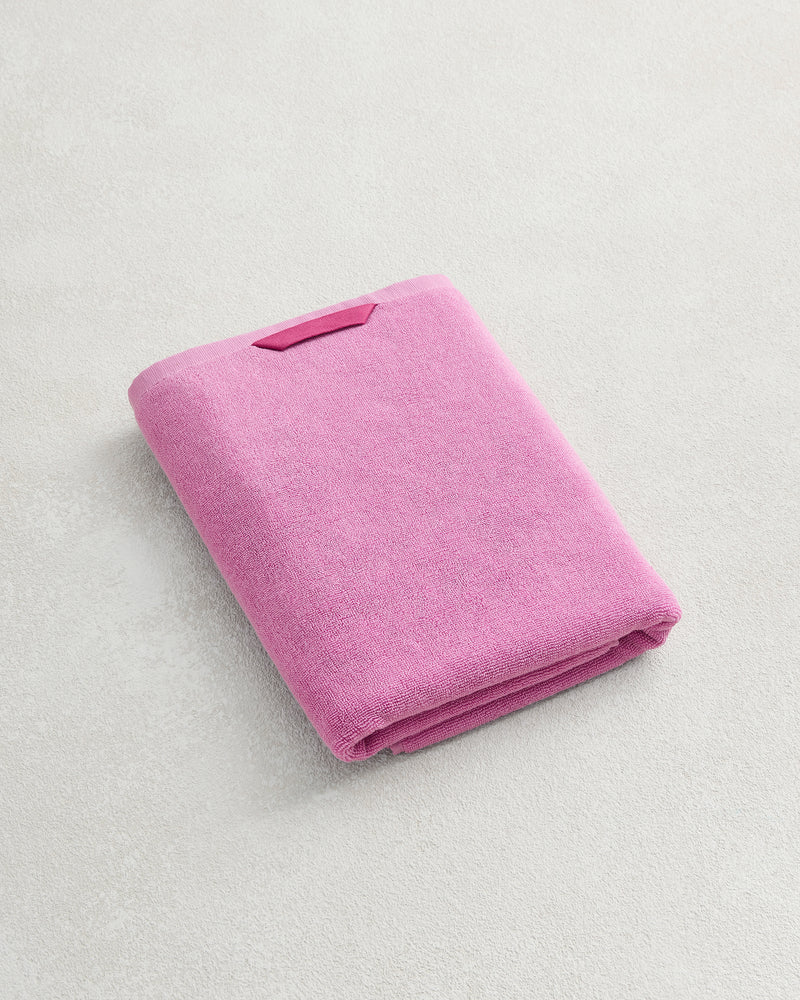 The Bed Threads Bubblegum 100% Cotton Terry Bath Towel, featuring a small rectangular tag and quick-drying 650GSM terry fabric, is folded on a light textured surface.