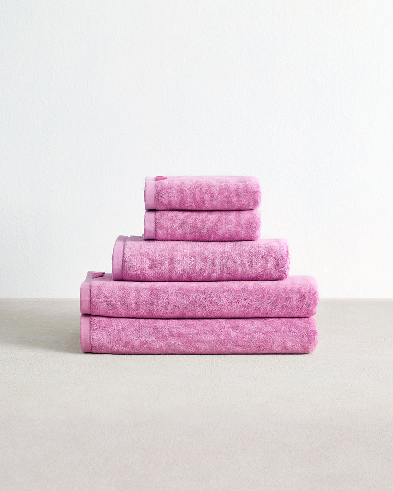 A set of four folded Bubblegum 100% Cotton Terry Bath Towels from Bed Threads rests neatly on a light beige surface against a plain white background.