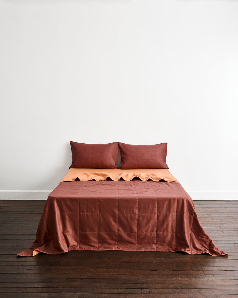 A bed dressed in a rust brown quilt cover in Cacao & Hazelnut with two matching pillowcases in Cacao, set against a plain white wall and dark wood floor.