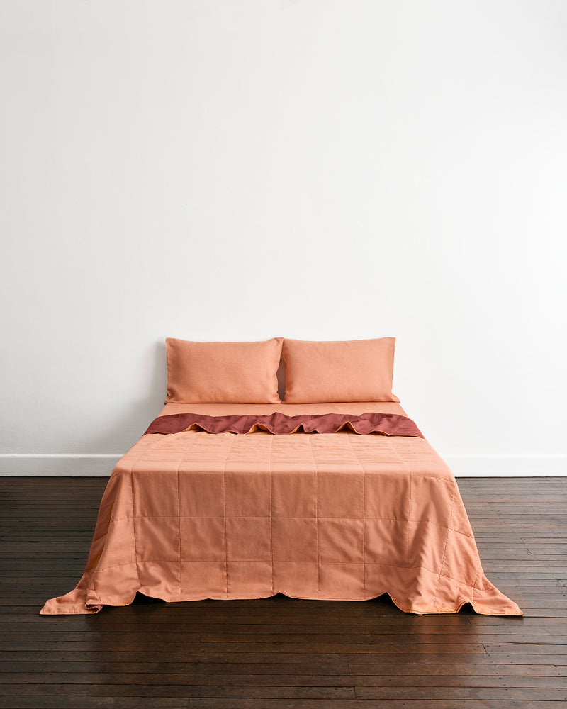 A bed dressed in a muted peach-orange quilt cover in Cacao & Hazelnut with two matching pillowcases in Hazelnut, set against a plain white wall and dark wood floor.
