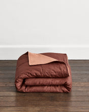 A folded brown quilt cover in Cacao & Hazelnut with or a Hazelnut underside, displayed on a dark wooden floor against a plain white wall. - view