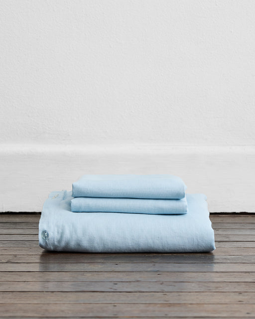 A neatly folded stack of light blue Coast 100% French Flax Linen Duvet Cover Set by Bed Threads sits on a wooden floor against a white wall, perfectly complementing your luxurious bedding ensemble.