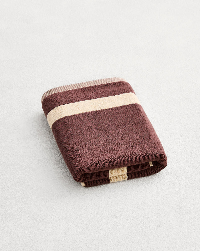 A folded cotton terry bath mat on a grey background, in a cacao colourway with a creme border. 