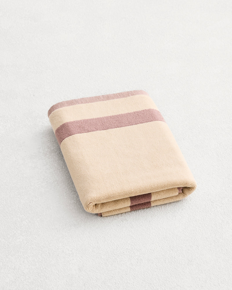 A folded cotton terry bath mat on a grey background, in a creme colourway with a lavender border. 