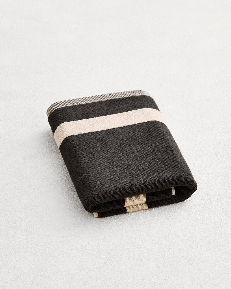 A folded cotton terry bath mat on a grey background, in a charcoal colourway with an oatmeal border. 