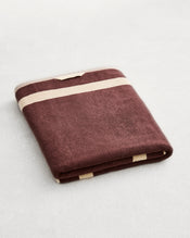 A folded cotton terry bath sheet on a grey background, in a cacao colourway with a creme border.  - view