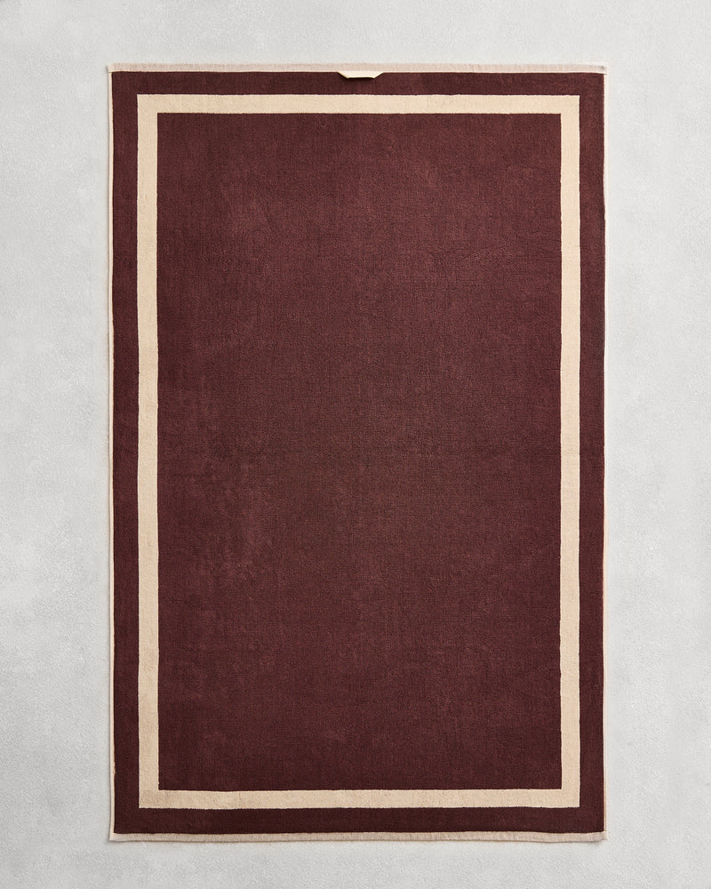 Flat cotton terry bath sheet on a grey background in a cacao colour with a creme border.