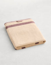 A folded cotton terry bath sheet on a grey background, in a creme colourway with a lavender border.  - view