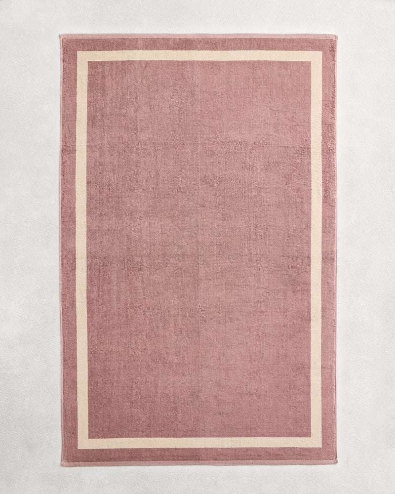 Flat cotton terry bath sheet on a grey background in a lavender colour with a creme border.