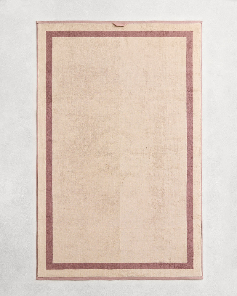 Flat cotton terry bath sheet on a grey background in a creme colour with a lavender border.