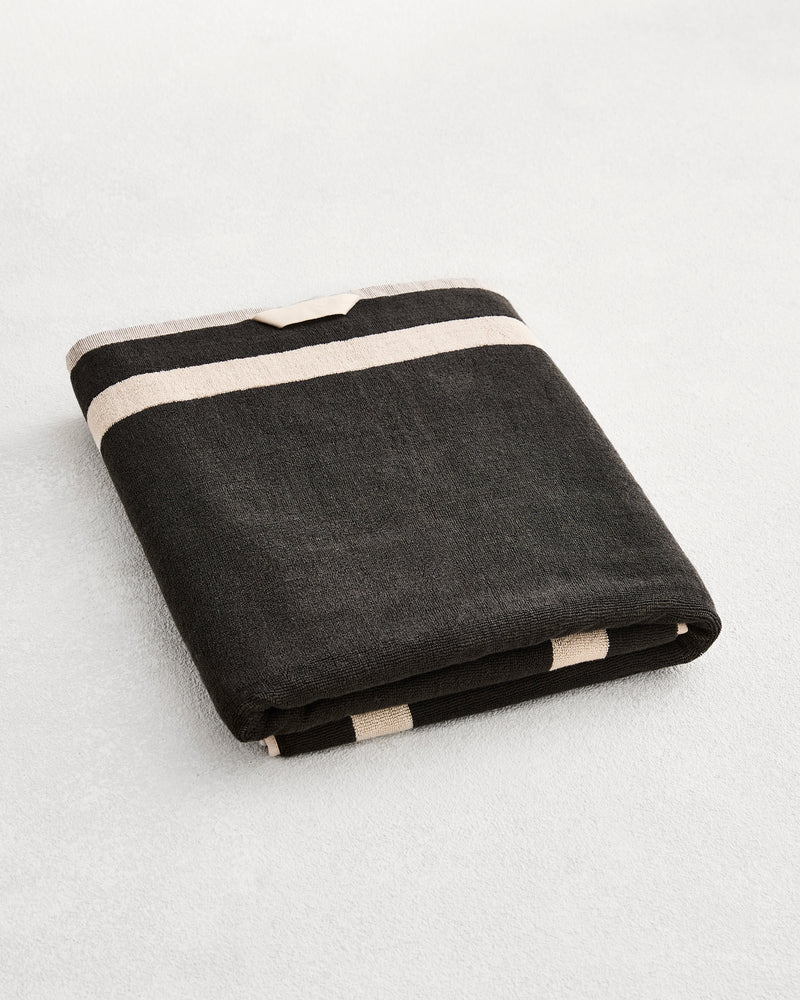A folded cotton terry bath sheet on a grey background, in a charcoal colourway with an oatmeal border. 