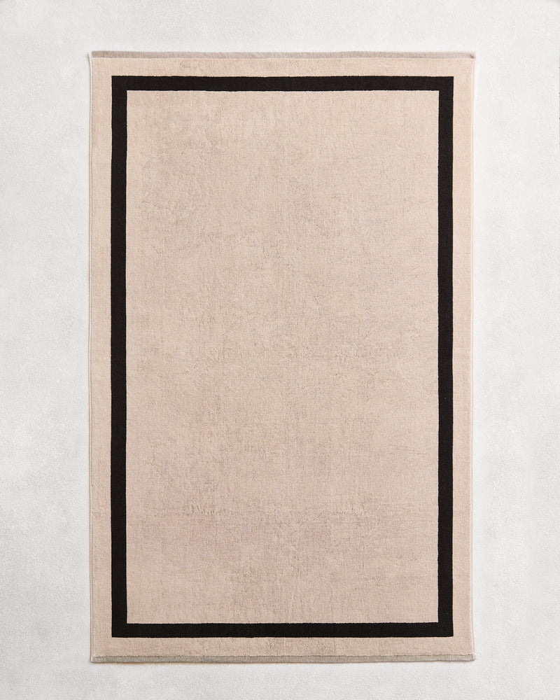 Flat cotton terry bath sheet on a grey background in an oatmeal colour with a charcoal border.