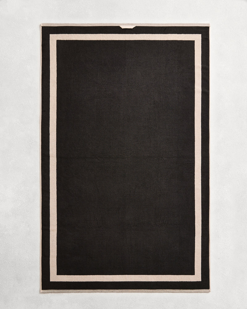 Flat cotton terry bath sheet on a grey background in a charcoal colour with an oatmeal border.