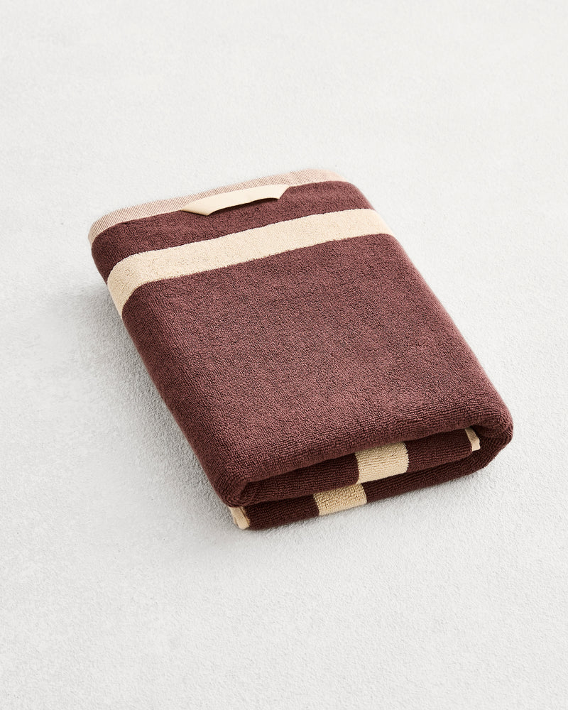 A folded cotton terry bath towel on a grey background, in a cacao colourway with a creme border. 