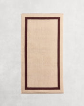 Flat cotton terry bath towel on a grey background in a creme colour with a cacao border. - view