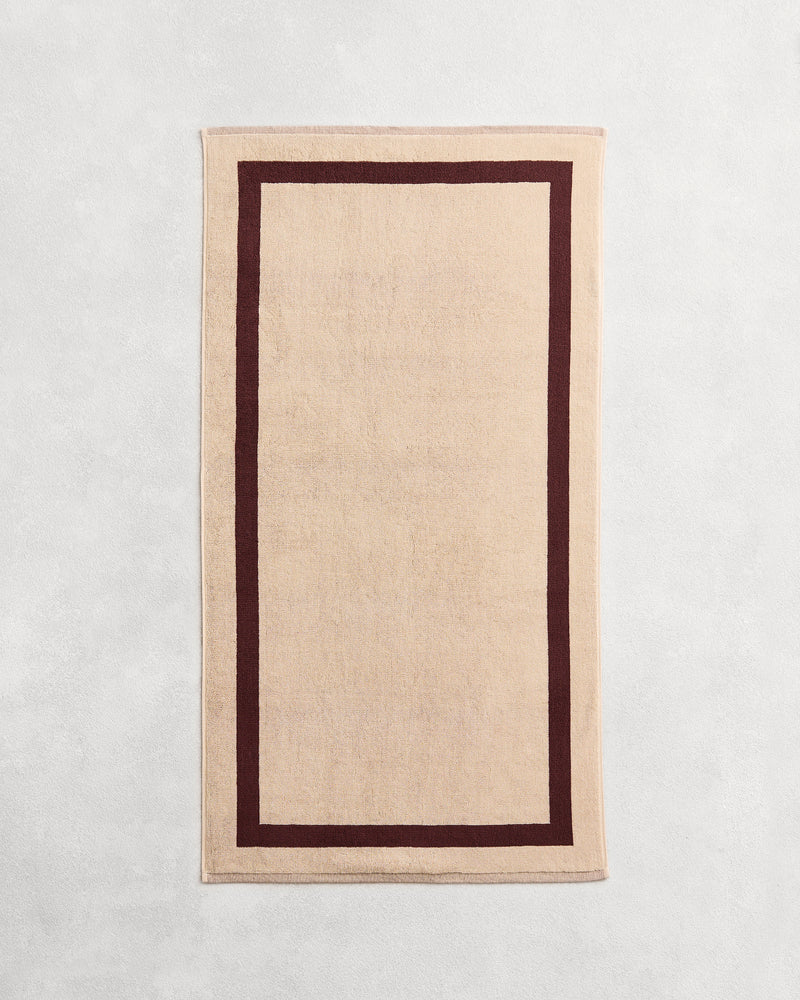 Flat cotton terry bath towel on a grey background in a creme colour with a cacao border.