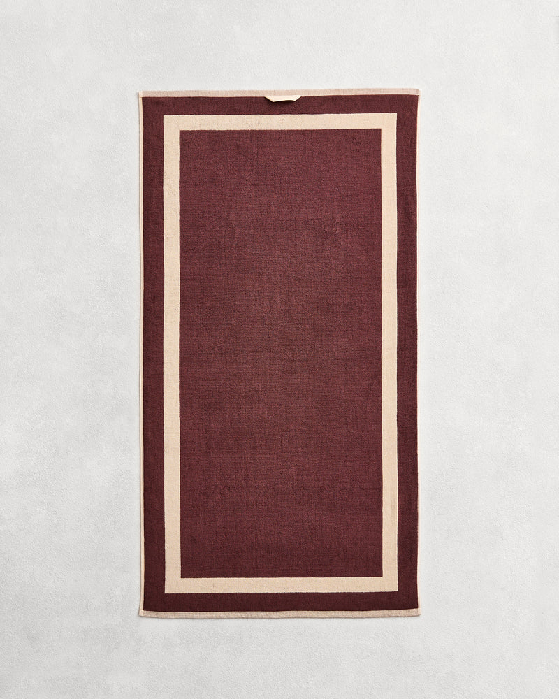 Flat cotton terry bath towel on a grey background in a cacao colour with a creme border.