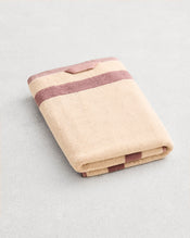 A folded cotton terry bath towel on a grey background, in a creme colourway with a lavender border.  - view